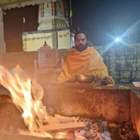 Bearded monk in orange robes sits by a large fire pit at night, holding a metal bowl, with a temple façade in the background.