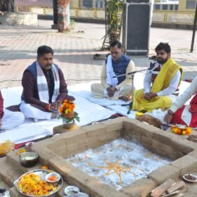 Group of men in traditional Indian attire sit on a white cloth around a square cremation pit, offering flowers as a lead speaker speaks into a microphone.