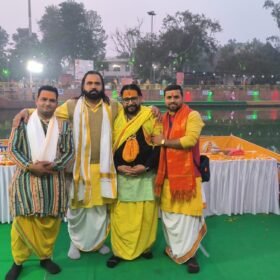 Four men in colorful traditional Indian clothing pose together at a festival by a decorated float and river, offering trays visible in the background.