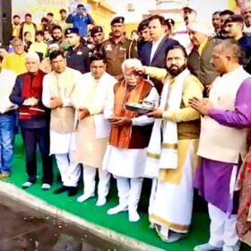 Group of Indian men in traditional clothing gather on a green carpet by a water body while a man in the center gets a blessing from another, as onlookers and police stand behind.