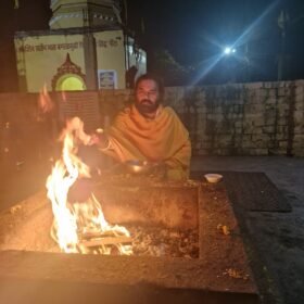 Bearded man in saffron robes sits beside a large fire in a stone outdoor hearth at night, with a temple wall in the background.