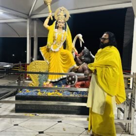 Statue of Goddess Durga in a bright yellow sari, holding a weapon, with a devotee in yellow performing a ritual nearby inside a sheltered shrine.