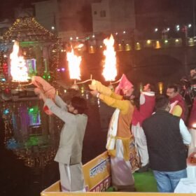 People on a boat performing a fire ritual on a river at night, with colorful lights reflecting on the water and a crowd nearby.