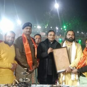 Group of men in colorful traditional attire posing around a ceremonial table with a framed document during a nighttime outdoor event by a lit water feature.