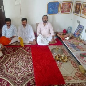 Three men sit cross-legged on festive red carpets during a Hindu ritual, with framed deities on the wall and a shrine area nearby.