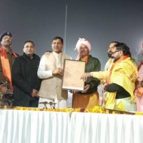 Group of men on a stage presenting a framed certificate during a ceremony, wearing traditional Indian attire and saffron scarves.