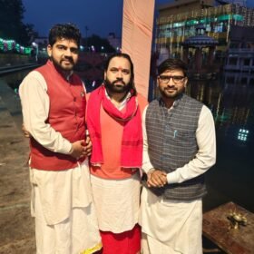Three men in traditional Indian attire posing together by a riverside or temple area at dusk, with festive lights in the background and red tilaks on their foreheads.