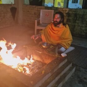 Bearded man in orange robes tending a fire in a stone pit of a courtyard, with a metal bowl nearby and temple walls in the background.