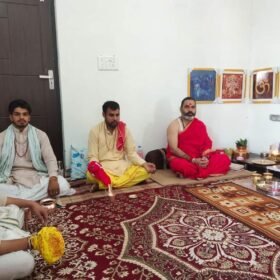 Four men sit cross-legged on a patterned rug in a room with a Hindu altar and deity images on the wall.