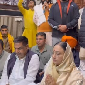 Group of people seated in a temple room as a man in orange holds a microphone and speaks, with religious portraits on the wall behind him.