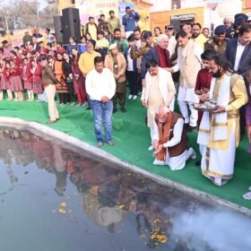 A ceremonial gathering by a pond with a priest bowing and making offerings on a green platform, surrounded by onlookers in traditional attire and schoolchildren in red uniforms on the left.