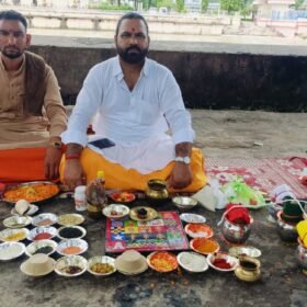 Two men sit cross‑legged on the ground performing a Hindu puja with bowls of colored powders, flowers, and ritual items laid out before them by the riverside.