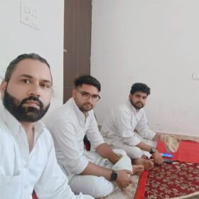Three men in white shirts sit on a red patterned rug indoors, facing the camera.