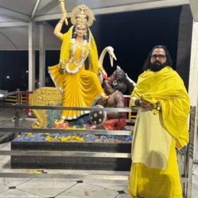 Hindu goddess Durga statue in a yellow sari lifting a golden mace on a decorated pedestal; a man in a bright yellow robe stands beside it inside a temple at night.