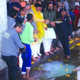 Group of men performing a religious rite at a temple ghats, standing in shallow water while a man in yellow robes pours water over others, with onlookers and security nearby.