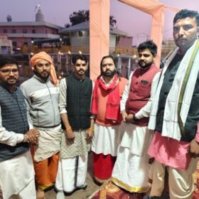 Group of six men in traditional Indian attire posing together near a water body at a temple area during a festival scene