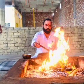 Man with beard and red tilak on forehead tending a ceremonial fire in a brick courtyard, using tongs from a metal bowl nearby.
