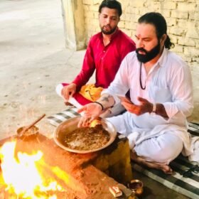 Two men perform a Hindu puja by a sacred fire, preparing offerings in a metal plate beside an orange flame.