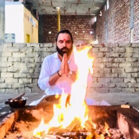 Bearded man in a white shirt sits cross-legged, hands pressed in prayer, beside a tall fire in a rectangular pit.